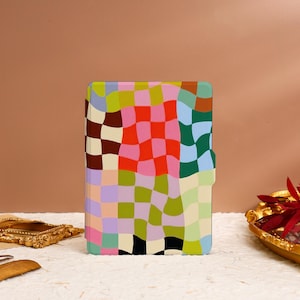 Pode incluir: Uma capa estampada com um design xadrez em tons de rosa, vermelho, verde, azul e castanho. A capa é retangular com cantos arredondados e fecho magnético. A capa está sobre uma superfície branca.