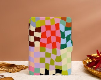 May include: A colorful, patterned case with a checkerboard design in shades of pink, red, green, blue, and brown. The case is rectangular with rounded corners and a magnetic closure. The case is sitting on a white surface.