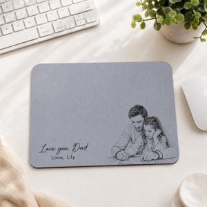 Op de afbeelding: Een rechthoekige grijze muismat met een potloodschets van een vader en kind. De tekst "Love you, Dad, Love, Lily" staat op de muismat. Een wit toetsenbord en muis staan op de achtergrond.
