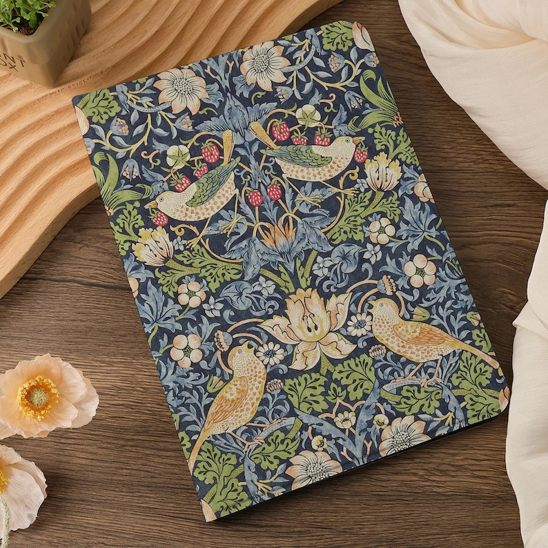 May include: A rectangular notebook with a navy blue floral pattern featuring birds, flowers, and berries. The design includes shades of green, cream, and red. The notebook is lying on a wooden surface.