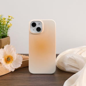 May include: A beige and orange gradient phone case. The case has a smooth finish and a camera cutout with three black lenses. The case is on a wooden surface with flowers and fabric in the background.