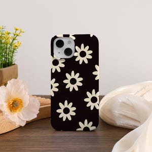 May include: A black phone case featuring a repeating pattern of cream-colored daisy flowers. The case has a glossy finish and is designed to fit a smartphone. The daisies are evenly spaced across the surface, creating a retro floral design.