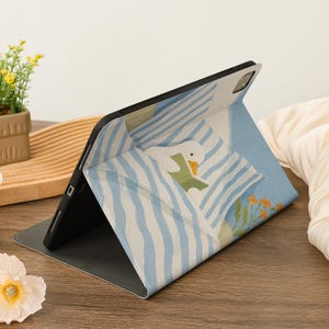 Puede incluir: Una funda para iPad con una ilustraci&oacute;n de un ganso leyendo en la cama. La funda presenta un patr&oacute;n de rayas azules y blancas, un marco negro y una base gris. El dise&ntilde;o incluye flores y un fondo azul claro.