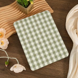 May include: A rectangular tablet case with a green and white gingham pattern. The case is resting on a wooden surface, with a small potted plant and fabric in the background. The case is a neutral color.