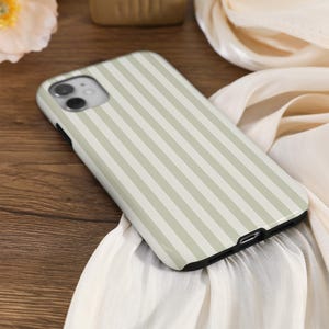 May include: A smartphone case with a vertical striped pattern in light green and white. The case has a black border and is resting on a wooden surface with a white fabric.