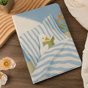 Puede incluir: Cuaderno con una ilustraci&oacute;n caprichosa de un pato leyendo un libro en la cama. La portada presenta un patr&oacute;n de rayas azules y blancas, un fondo azul claro y un peque&ntilde;o ramo de flores naranjas.