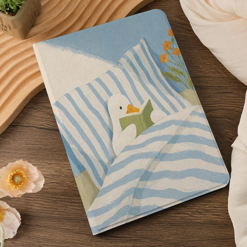 Puede incluir: Cuaderno con una ilustraci&oacute;n caprichosa de un pato leyendo un libro en la cama. La portada presenta un patr&oacute;n de rayas azules y blancas, un fondo azul claro y un peque&ntilde;o ramo de flores naranjas.