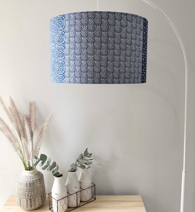 May include: A white lampshade with a blue and white geometric pattern. The lampshade is hanging from a white lamp stand. The lamp is in a room with a white wall and a wooden table with three vases on it.
