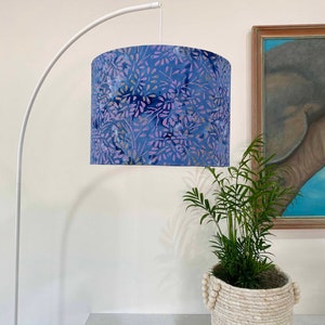 Pu&ograve; includere: Una lampada da terra ad arco bianca con un paralume a motivi floreali blu e viola. La lampada si trova in una stanza con una parete bianca e una pianta in un cesto intrecciato.