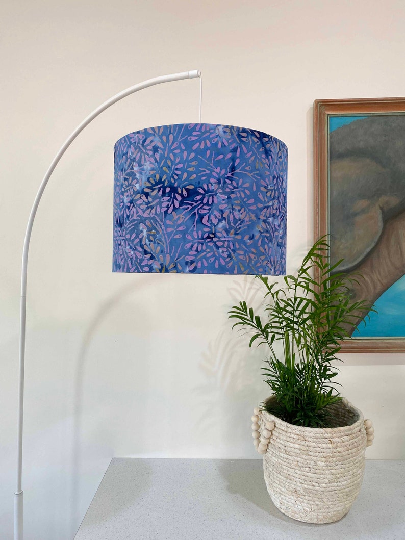 Pu&ograve; includere: Una lampada da terra ad arco bianca con un paralume a motivi floreali blu e viola. La lampada si trova in una stanza con una parete bianca e una pianta in un cesto intrecciato.