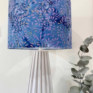 Pu&ograve; includere: Una lampada bianca con un paralume a motivi floreali blu e viola. La lampada ha una base in legno bianco con doghe verticali.