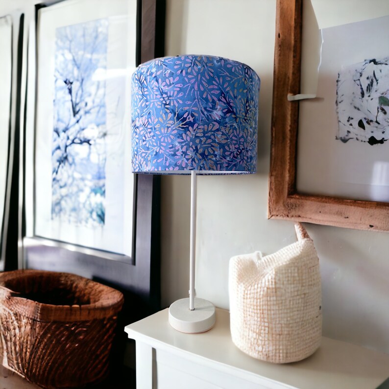 Pu&ograve; includere: Una lampada da tavolo bianca con un paralume a motivi floreali blu e viola. La lampada &egrave; su un tavolo bianco accanto a un cesto bianco intrecciato.