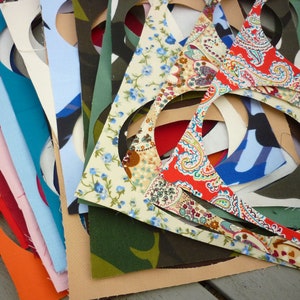 May include: A stack of fabric scraps in various colours and patterns, including floral, paisley, camouflage, and plain colours. The fabric scraps have been cut into circles with the centres removed.