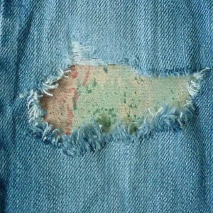 May include: Close-up of a blue denim fabric with a large, frayed hole revealing a faded, multi-coloured fabric underneath.