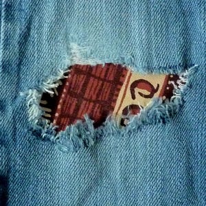 May include: A close-up of a pair of blue denim jeans with a large tear in the fabric. The tear is patched with a colourful fabric with a geometric pattern.
