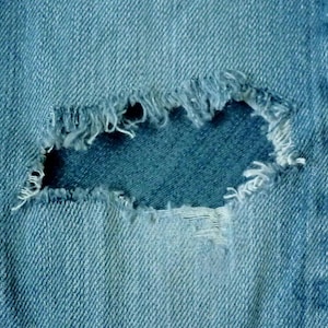 May include: Close-up of a blue denim fabric with a large hole in the center. The hole is frayed and has a rough texture.
