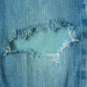 Könnte beinhalten: Nahaufnahme eines blauen Jeansstoffes mit einem ausgefransten Loch. Das Loch hat ausgefranste Kanten und ist ein häufiges Merkmal von Vintage-Jeans.