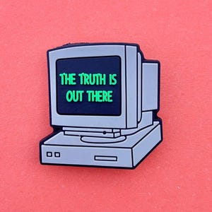 May include: A computer-shaped charm with the green text "THE TRUTH IS OUT THERE" on a black screen. The charm is mainly grey and black, with a retro design. The background is a solid coral colour.