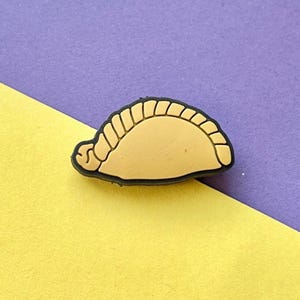 May include: A yellow and black rubber charm shaped like a pastry. The pastry has a crimped edge and a brown filling.
