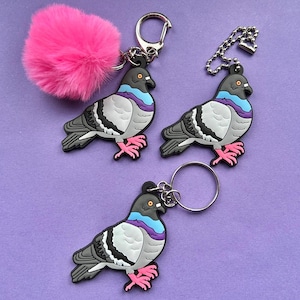 May include: Keyrings with a pigeon design in grey, white, black, blue, purple, and pink. One keyring has a pink pom-pom. The keyrings have silver-coloured metal rings and chains. The background is a solid purple colour.