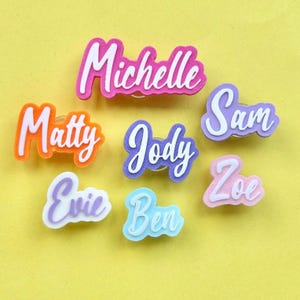 May include: A collection of seven custom name pins featuring layered acrylic designs. The names Michelle, Matty, Jody, Sam, Evie, Ben, and Zoe are displayed in white script lettering against colorful, solid-colored acrylic backings in pink, orange, purple, and blue.
