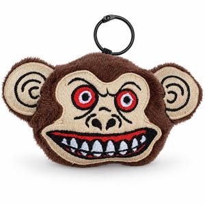 May include: A plush monkey keyring featuring a brown fuzzy head with large beige ears. The face has an embroidered design with red eyes, a black nose, and a wide mouth showing white teeth against a red background. A black metal ring is attached to the top.