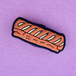 May include: A shoe charm in the shape of a sandwich. The charm has light orange bread with white and black details, outlined in dark blue. The charm is set against a purple background.