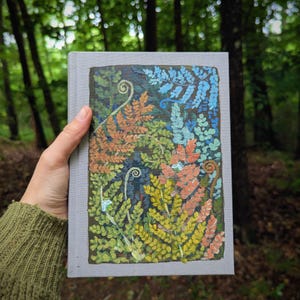 May include: A hand holds a book with a painted fern design. The cover features a detailed illustration of ferns in shades of orange, yellow, blue, and green against a dark background. The book's cover is a light gray color.