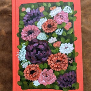 May include: A rectangular, coral-colored canvas with a floral painting. The painting features a variety of flowers in shades of pink, purple, orange, and white, surrounded by green leaves. The artwork is signed in the bottom right corner.