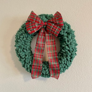 May include: A green yarn wreath with a red and white plaid bow. The bow is tied in a knot at the center and the ends of the bow are hanging down.
