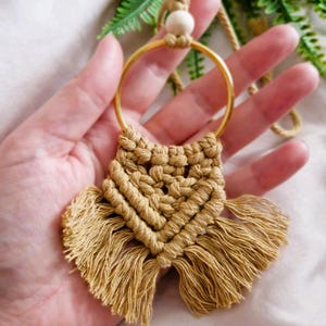 May include: A tan macrame necklace featuring a gold-coloured metal ring and a wooden bead. The pendant has a woven design with a fringe. The necklace is handmade.