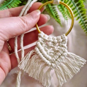 May include: A cream-coloured macrame wall hanging, featuring a gold metal ring and woven cotton cord. The design includes a fringed bottom. The piece is held by a hand, highlighting the texture and craftsmanship of the fibre art, suitable for home decor.