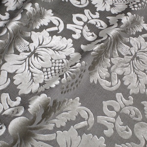 May include: Close-up of a silver fabric with a raised damask pattern. The fabric has a subtle sheen and a soft, luxurious feel.