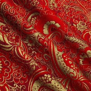 Op de afbeelding: Rode stof met een gouden paisley-patroon. De stof heeft een textuur en is geweven.