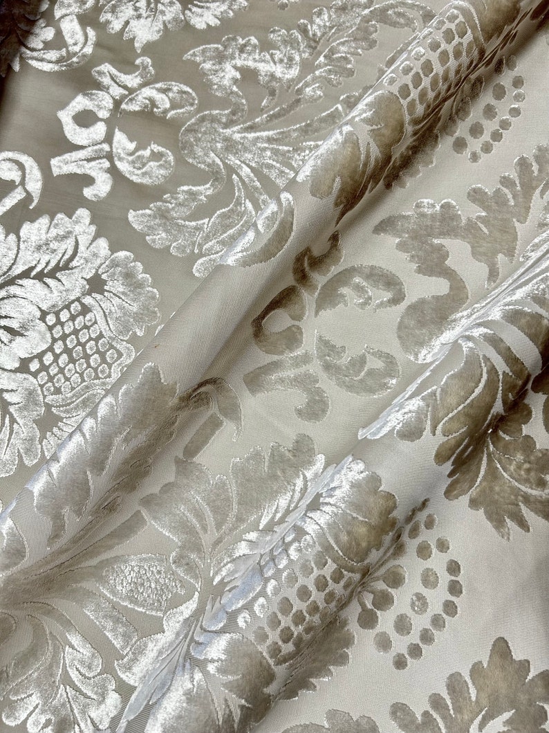 May include: A close-up of a white fabric with a raised, embossed pattern of silver-toned floral designs. The fabric has a soft, velvety texture.