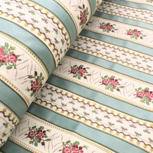 Op de afbeelding: Close-up van een stoffen rol met een herhalend patroon. Het ontwerp omvat afwisselende strepen in lichtblauw en gebroken wit. De gebroken witte strepen zijn versierd met bloemen- en bladmotieven in rood, groen en geel.