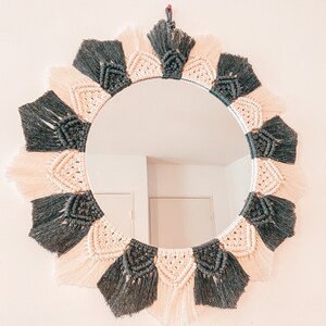 Peut inclure: Un miroir rond avec un cadre en macramé noir et crème. Le cadre est fait de cordons noués avec des détails en franges.