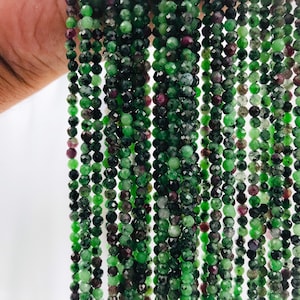 May include: A strand of green and pink faceted gemstone beads. The beads are small and round, and they are strung on a clear thread. The beads are all different shades of green and pink, and they have a natural, unpolished look.