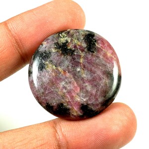 May include: A round, polished cabochon of pink and black ruby with gold flecks. The stone is held in a hand.