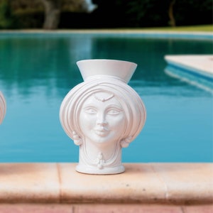 May include: White ceramic vase shaped like a woman's head with a turban. The vase has a smooth, glossy finish and is sitting on a stone surface.