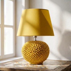 Op de afbeelding: Een gele tafellamp met een getextureerde, bolvormige voet en een bijpassende lampenkap. De lamp heeft een zilverkleurige steel. De lamp staat op een tafel met een patroon. De lamp is ongeveer 46 cm hoog.