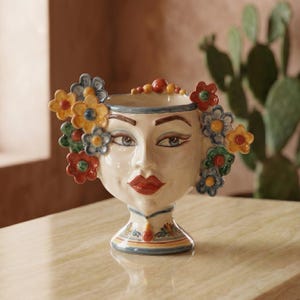 May include: A ceramic head-shaped vase featuring a painted face with red lips and dark eyebrows. The vase is adorned with three-dimensional colorful flowers in shades of blue, yellow, green, and red around the sides and a small red bead at the base.