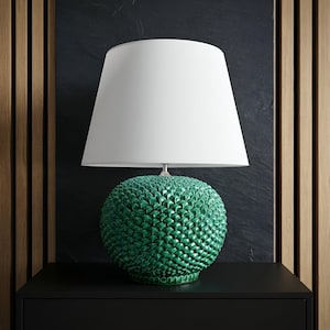 Puede incluir: Una lámpara de mesa de cerámica verde con una pantalla blanca. La base de la lámpara tiene un patrón texturizado en forma de escamas. La lámpara está sobre una superficie negra contra un fondo oscuro con paneles de madera.