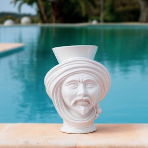 May include: White ceramic vase shaped like a man's head wearing a turban. The vase has a simple design and is perfect for adding a touch of modern style to any room.