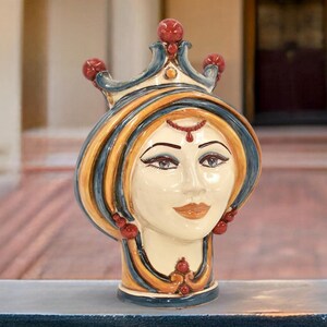 May include: A ceramic bust of a woman with a crown. The face is cream-colored with detailed features, including dark eyes and red accents. The crown and hair are adorned with blue, yellow, and red details. The bust is a decorative item.