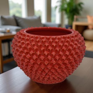 May include: A round, red ceramic decorative bowl with a textured, scale-like pattern covering its exterior. The bowl sits on a dark wooden surface, with a blurred background of a living room.