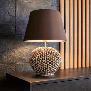 May include: A decorative table lamp with a brown lampshade and a textured, spherical base. The base has a pattern of small, overlapping shapes in shades of beige and brown. The lamp is set on a dark surface.