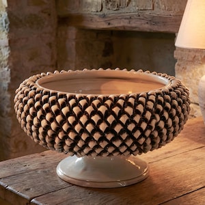 Może przedstawiać: Ozdobna, beżowa ceramiczna miska z teksturowanym, łuskowatym wzorem na zewnątrz. Misa stoi na podstawie i jest prezentowana na drewnianej powierzchni. Wnętrze miski jest gładkie i jasne. Średnica miski wynosi około 30 cm.