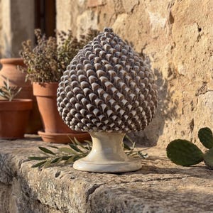 May include: A ceramic pinecone sculpture with a textured surface, in shades of brown and cream. The sculpture sits on a cream-coloured pedestal base. The background includes terracotta pots with plants and a stone wall.