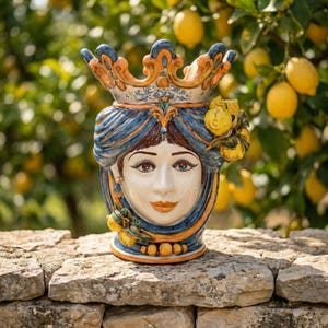 May include: A ceramic vase with a woman's face, adorned with a crown and blue and orange accents. The vase features a detailed face with brown hair and is decorated with lemons. The vase is placed on a stone wall with a lemon tree in the background.